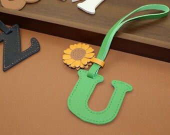May include: A green leather letter "U" with a sunflower accent and a green strap. The letter is stitched with white thread. Other leather letters are visible in the background.