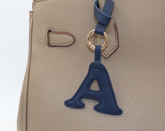 May include: A taupe-colored handbag with a textured surface featuring a dark blue leather letter A charm. The charm is attached to the bag handle by a gold-toned metal ring and a dark blue leather strap, creating a distinct contrast in color.