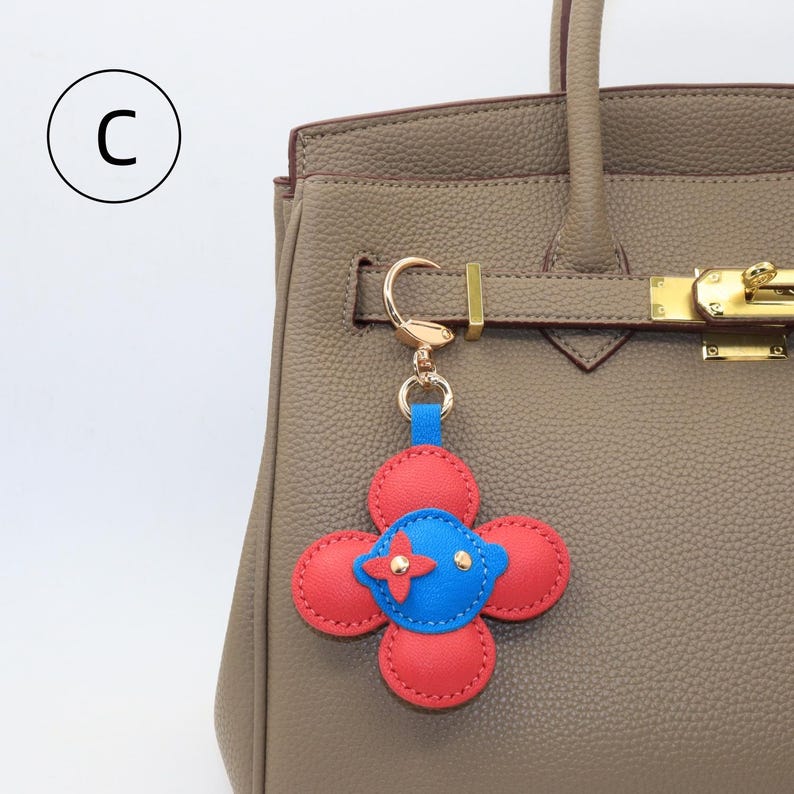 K&ouml;nnte beinhalten: Ein rot-blauer, bl&uuml;tenf&ouml;rmiger Schl&uuml;sselanh&auml;nger mit goldfarbener Hardware ist an einer taupefarbenen Lederhandtasche befestigt. Der Schl&uuml;sselanh&auml;nger hat eine blaue Mitte mit einem roten Kreuz und zwei goldfarbenen Nieten. Die Handtasche hat einen goldenen Verschluss und Griff.