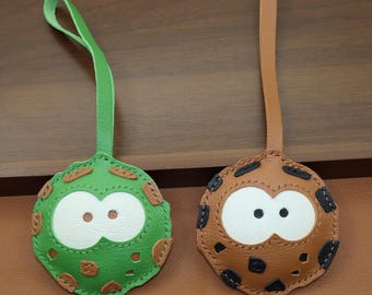 May include: Two leather keychains with cartoon-like faces. One is green with white eyes and brown spots, the other is brown with white eyes and black spots. Both have matching leather loops for attachment.