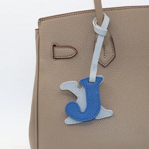 May include: Close-up of a beige leather handbag with a light blue and blue leather letter "J" charm. The handbag has a top handle and a clasp closure. The charm is attached to the handle with a light blue leather strap.