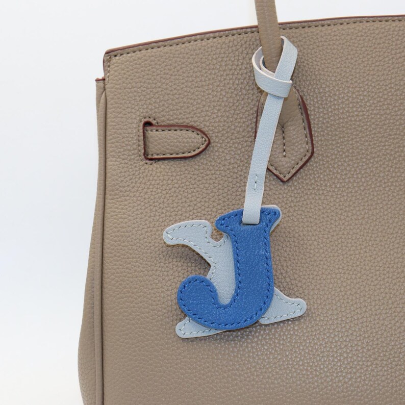 May include: Close-up of a beige leather handbag with a light blue and blue leather letter "J" charm. The handbag has a top handle and a clasp closure. The charm is attached to the handle with a light blue leather strap.
