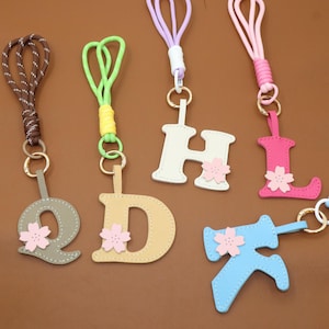 May include: A collection of colorful keychains featuring letters of the alphabet. Each letter is a different color, such as gray, beige, white, pink, and blue, and has a small pink flower attached. The keychains have various colored cords.