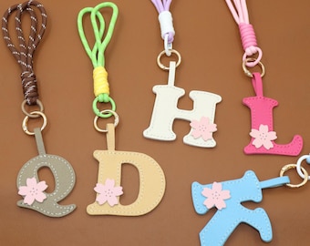 May include: A collection of colorful keychains featuring letters of the alphabet. Each letter is a different color, such as gray, beige, white, pink, and blue, and has a small pink flower attached. The keychains have various colored cords.