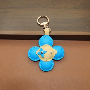 K&ouml;nnte beinhalten: Ein Leder-Schl&uuml;sselanh&auml;nger in Form eines vierbl&auml;ttrigen Kleeblatts. Das Kleeblatt hat hellblaue Bl&uuml;tenbl&auml;tter mit wei&szlig;en N&auml;hten und eine beige Mitte mit einer kleinen blauen Blume und goldenen Akzenten. Der Schl&uuml;sselanh&auml;nger hat einen goldenen Ring und einen beigefarbenen Lederriemen.