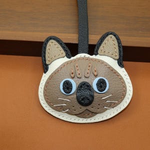 May include: A Siamese cat-shaped leather keyring with a black strap. The cat's face features blue eyes, a black nose, and brown and white details. The keyring is stitched with a contrasting thread.