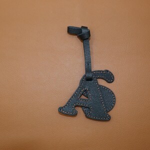 May include: A dark teal leather letter "A" luggage tag with tan stitching. The tag is attached to a strap with a knotted end. The background is a solid tan color.