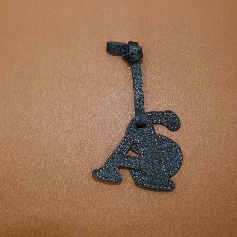 May include: A dark teal leather letter "A" luggage tag with tan stitching. The tag is attached to a strap with a knotted end. The background is a solid tan color.