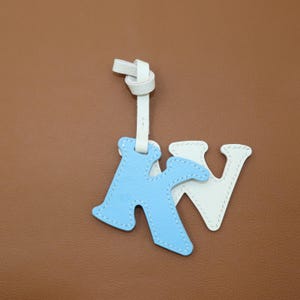 May include: A leather keychain featuring the letters "K" in light blue and "V" in white, stitched with white thread. The keychain has a white leather strap and a knot at the top. The background is a solid brown color.