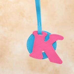 May include: A pink leather letter "K" charm, stitched onto a blue circular base, with a matching blue strap for hanging. The charm is suspended against a neutral background.