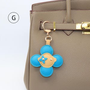 K&ouml;nnte beinhalten: Ein blauer und beigefarbener Lederblumen-Schl&uuml;sselanh&auml;nger mit einem goldfarbenen Verschluss ist an einer taupefarbenen Handtasche befestigt. Der Schl&uuml;sselanh&auml;nger hat ein vierbl&auml;ttriges Design mit einer kleinen Blume und zwei goldfarbenen Nieten in der Mitte.