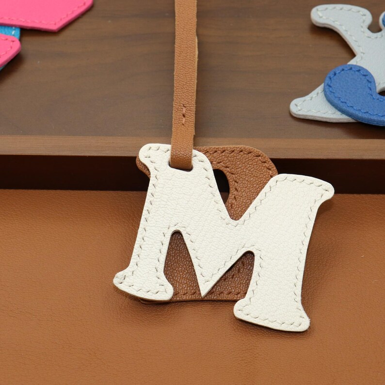 May include: A leather bag charm in the shape of the letter "M". The charm is white with brown backing and a brown leather strap. The charm is stitched with white thread. The background is a brown leather surface.