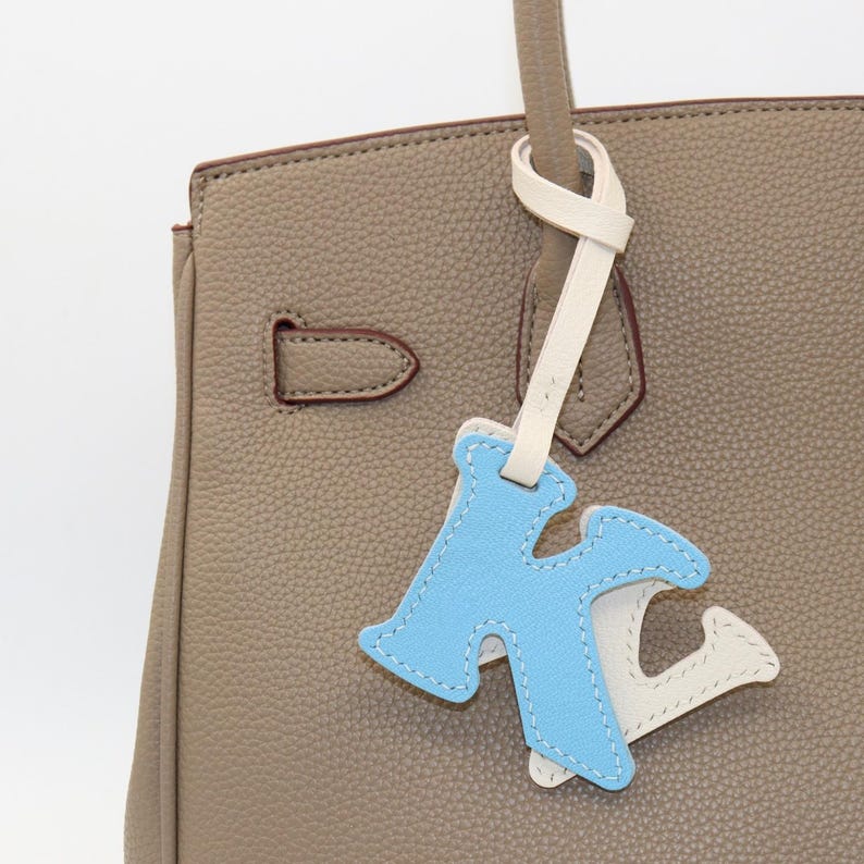 May include: A light blue and white leather letter "K" bag charm with white stitching, hanging from a taupe handbag. The handbag has a top handle and a silver-colored clasp.