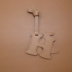 May include: A tan leather bag charm in the shape of the letter "H". The charm is attached to a matching leather strap with a knotted detail at the top. The background is a solid tan color.