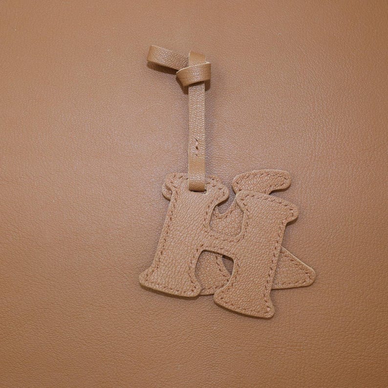 May include: A tan leather bag charm in the shape of the letter "H". The charm is attached to a matching leather strap with a knotted detail at the top. The background is a solid tan color.