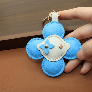 K&ouml;nnte beinhalten: Ein blauer und wei&szlig;er Leder-Schl&uuml;sselanh&auml;nger in Blumenform mit goldfarbener Hardware. Der Schl&uuml;sselanh&auml;nger hat ein vierbl&auml;ttriges Design mit hellblauem &Auml;u&szlig;eren und einer wei&szlig;en Mitte. Die Mitte hat ein goldenes Kreuz-Design und zwei kleine goldene Nieten.