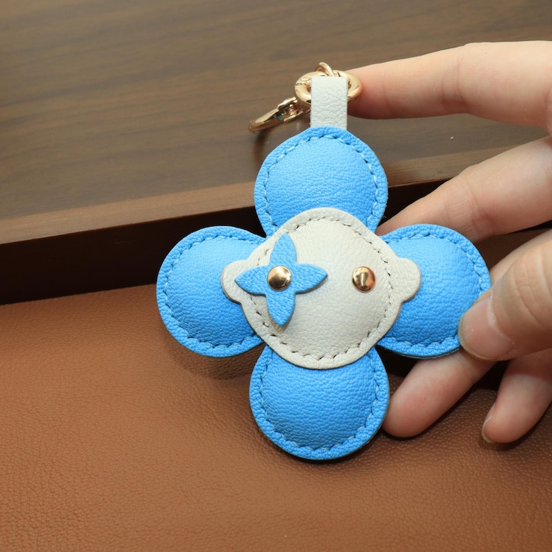 K&ouml;nnte beinhalten: Ein blauer und wei&szlig;er Leder-Schl&uuml;sselanh&auml;nger in Blumenform mit goldfarbener Hardware. Der Schl&uuml;sselanh&auml;nger hat ein vierbl&auml;ttriges Design mit hellblauem &Auml;u&szlig;eren und einer wei&szlig;en Mitte. Die Mitte hat ein goldenes Kreuz-Design und zwei kleine goldene Nieten.