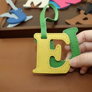May include: A yellow and green leather letter "E" with a green strap. The letter is stitched with yellow thread. Other leather shapes are visible in the background, including a blue guitar and other colorful shapes.