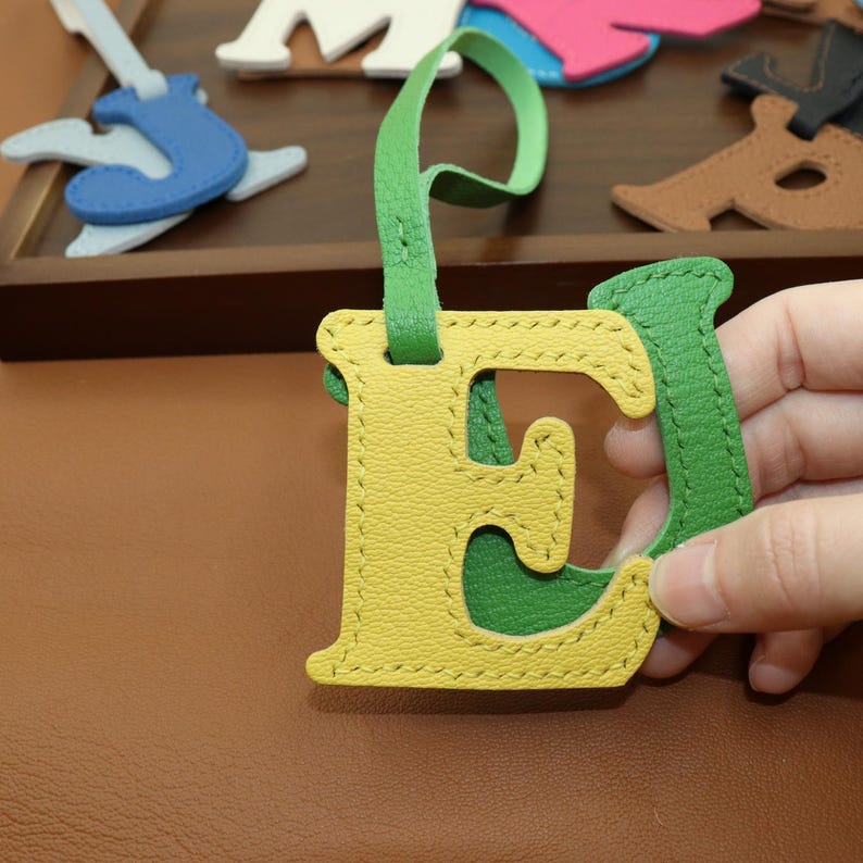 May include: A yellow and green leather letter "E" with a green strap. The letter is stitched with yellow thread. Other leather shapes are visible in the background, including a blue guitar and other colorful shapes.