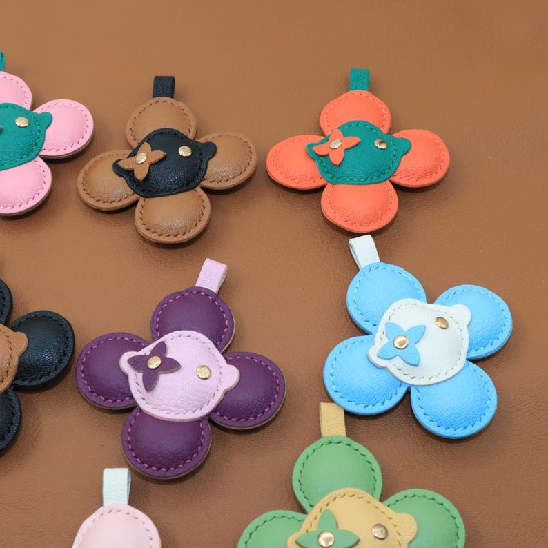 K&ouml;nnte beinhalten: Eine Sammlung von ledernen, bl&uuml;tenf&ouml;rmigen Anh&auml;ngern. Jeder Anh&auml;nger hat ein vierbl&auml;ttriges Design mit einem kontrastierenden Mittelteil und einer kleinen Schlaufe zur Befestigung. Farben sind Rosa, Orange, Gr&uuml;n, Blau, Lila, Schwarz und Braun.