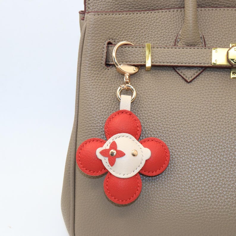 K&ouml;nnte beinhalten: Ein roter und wei&szlig;er Leder-Bl&uuml;ten-Schl&uuml;sselanh&auml;nger mit goldfarbener Hardware. Der Anh&auml;nger ist an einer grauen Lederhandtasche befestigt. Die Blume hat vier rote Bl&uuml;tenbl&auml;tter und eine wei&szlig;e Mitte mit rotem Kreuzmuster.