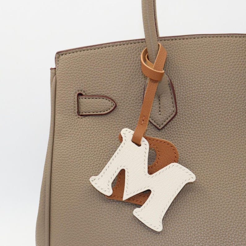 May include: Close-up of a taupe leather handbag with a matching leather handle. A decorative leather tag in the shape of the letter "M" is attached to the handle. The tag is white with brown accents and stitched edges.