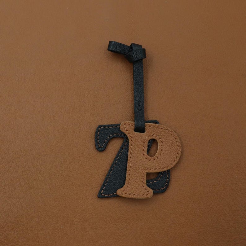 May include: A leather bag charm featuring a dark navy blue letter Z layered behind a tan letter P. The letters are crafted from textured leather with visible stitching along the edges and are suspended from a matching dark navy blue leather strap.