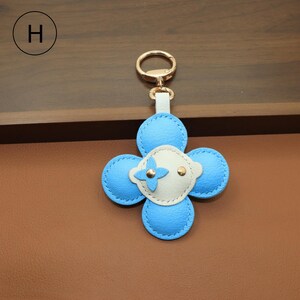 K&ouml;nnte beinhalten: Ein blau-wei&szlig;er Leder-Schl&uuml;sselanh&auml;nger in Form einer vierbl&auml;ttrigen Blume. Der Schl&uuml;sselanh&auml;nger hat einen goldfarbenen Verschluss und einen kleinen wei&szlig;en Lederriemen. Die Blume hat einen goldfarbenen Akzent in der Mitte.