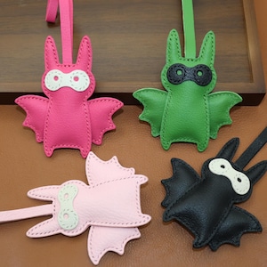 Peut inclure: Quatre porte-clés en cuir en forme de chauve-souris, roses, verts, rose clair et noirs. Chaque chauve-souris a un design cousu avec des détails d'yeux blancs ou noirs et une sangle assortie. Les chauves-souris ont des ailes déployées.