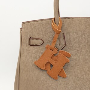 May include: Close-up of a taupe leather handbag with a matching handle and a decorative leather charm. The charm is in the shape of the letter "H" in a lighter shade of brown. The bag has a structured design.