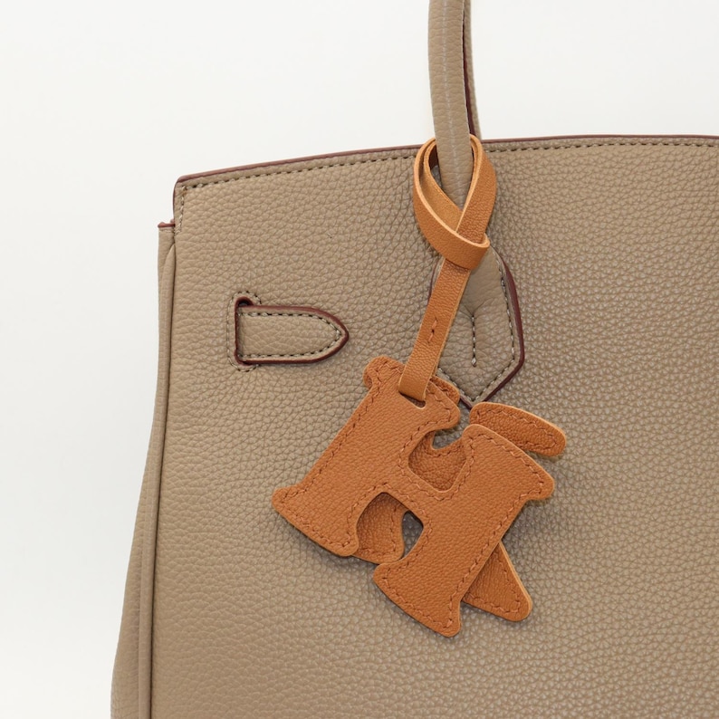 May include: Close-up of a taupe leather handbag with a matching handle and a decorative leather charm. The charm is in the shape of the letter "H" in a lighter shade of brown. The bag has a structured design.