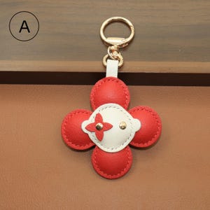 K&ouml;nnte beinhalten: Ein roter und wei&szlig;er Leder-Schl&uuml;sselanh&auml;nger in einem vierbl&auml;ttrigen Kleeblatt-Design. Das Kleeblatt hat vier rote Bl&uuml;tenbl&auml;tter und eine wei&szlig;e Mitte mit goldenem Akzent. Der Schl&uuml;sselanh&auml;nger hat einen goldfarbenen Verschluss.