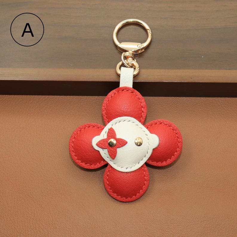 K&ouml;nnte beinhalten: Ein roter und wei&szlig;er Leder-Schl&uuml;sselanh&auml;nger in einem vierbl&auml;ttrigen Kleeblatt-Design. Das Kleeblatt hat vier rote Bl&uuml;tenbl&auml;tter und eine wei&szlig;e Mitte mit goldenem Akzent. Der Schl&uuml;sselanh&auml;nger hat einen goldfarbenen Verschluss.