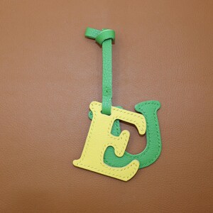 May include: A leather bag charm featuring the letters "E" and "J" in yellow and green. The "E" is yellow with green stitching, and the "J" is green with yellow stitching. A green strap is attached to the top, with a knot at the end.
