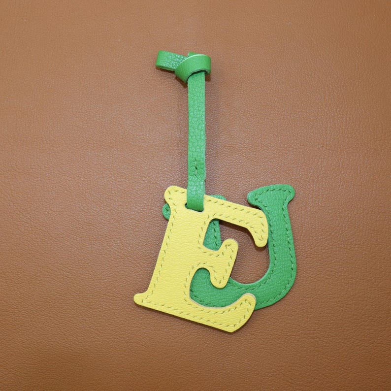 May include: A leather bag charm featuring the letters "E" and "J" in yellow and green. The "E" is yellow with green stitching, and the "J" is green with yellow stitching. A green strap is attached to the top, with a knot at the end.