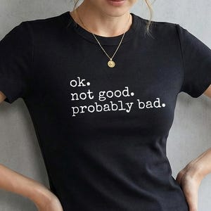 May include: Black crew neck t-shirt with the text "ok. not good. probably bad." in white. The shirt is paired with blue jeans. A woman is wearing the shirt and a gold necklace.
