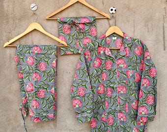May include: A pajama set consisting of a long-sleeved button-down shirt and matching pants, both featuring a gray background with a repeating pattern of pink flowers and green leaves. The set includes a small matching fabric pouch with a drawstring.