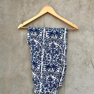 May include: A pair of trousers featuring a white base with a repeating dark blue floral and botanical print. The trousers are folded over a light-colored wooden clothes hanger, which is suspended from a small round white button labeled HOMERUN.