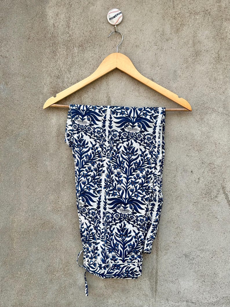 May include: A pair of trousers featuring a white base with a repeating dark blue floral and botanical print. The trousers are folded over a light-colored wooden clothes hanger, which is suspended from a small round white button labeled HOMERUN.