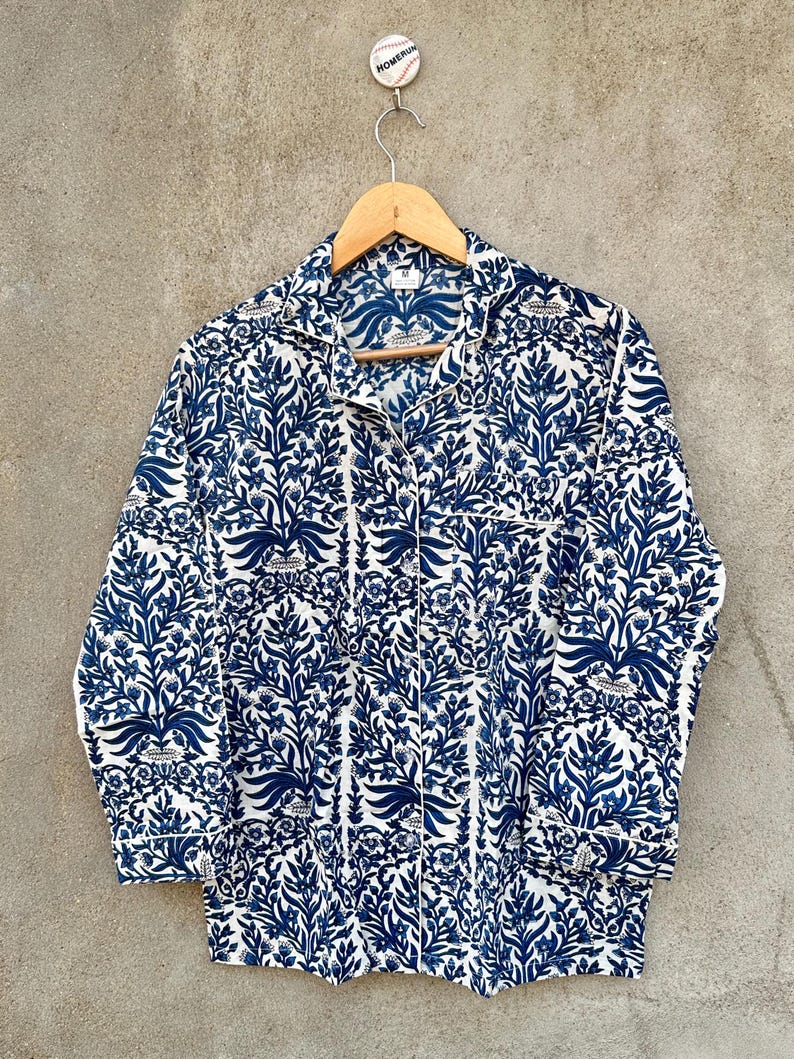 May include: A long-sleeve button-down shirt featuring a repeating dark blue floral and botanical pattern on a white background. The shirt has a notched collar, a single chest pocket, and white piping trim along the collar, pocket, and cuffs.