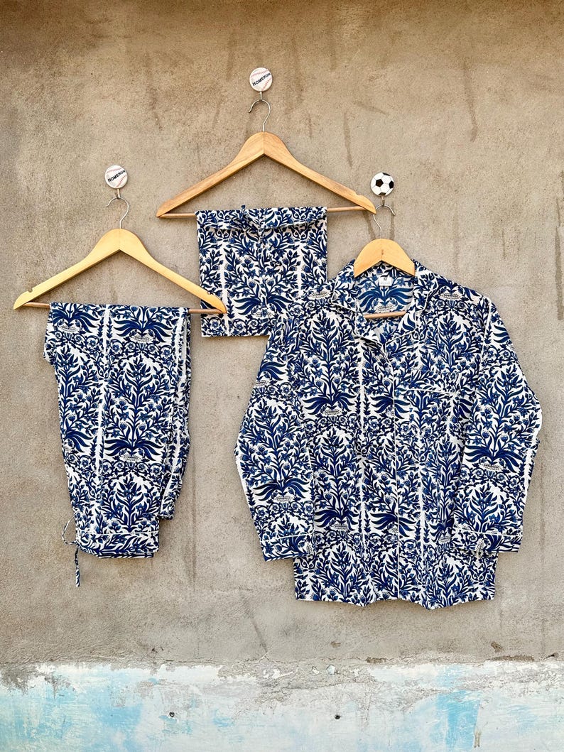 May include: A pajama set consisting of a long-sleeved button-down shirt and matching pants, featuring a repeating blue and white floral botanical print. The garments are displayed on wooden hangers against a textured gray wall surface.