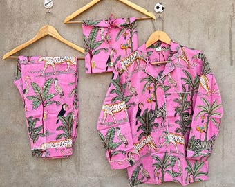 May include: A pink pajama set consisting of a long-sleeved button-down shirt and matching pants, accompanied by a small drawstring bag. The fabric features a repeating jungle print with leopards, zebras, toucans, and palm trees in green and yellow.