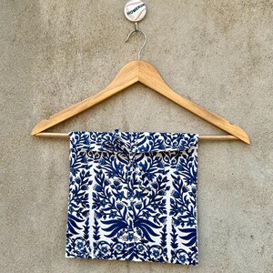May include: A square fabric pouch featuring a blue and white floral pattern, suspended from a wooden hanger. The pouch has a tie closure at the top center. Above the hanger, a small circular pin displays the word HOMERUN in black text.