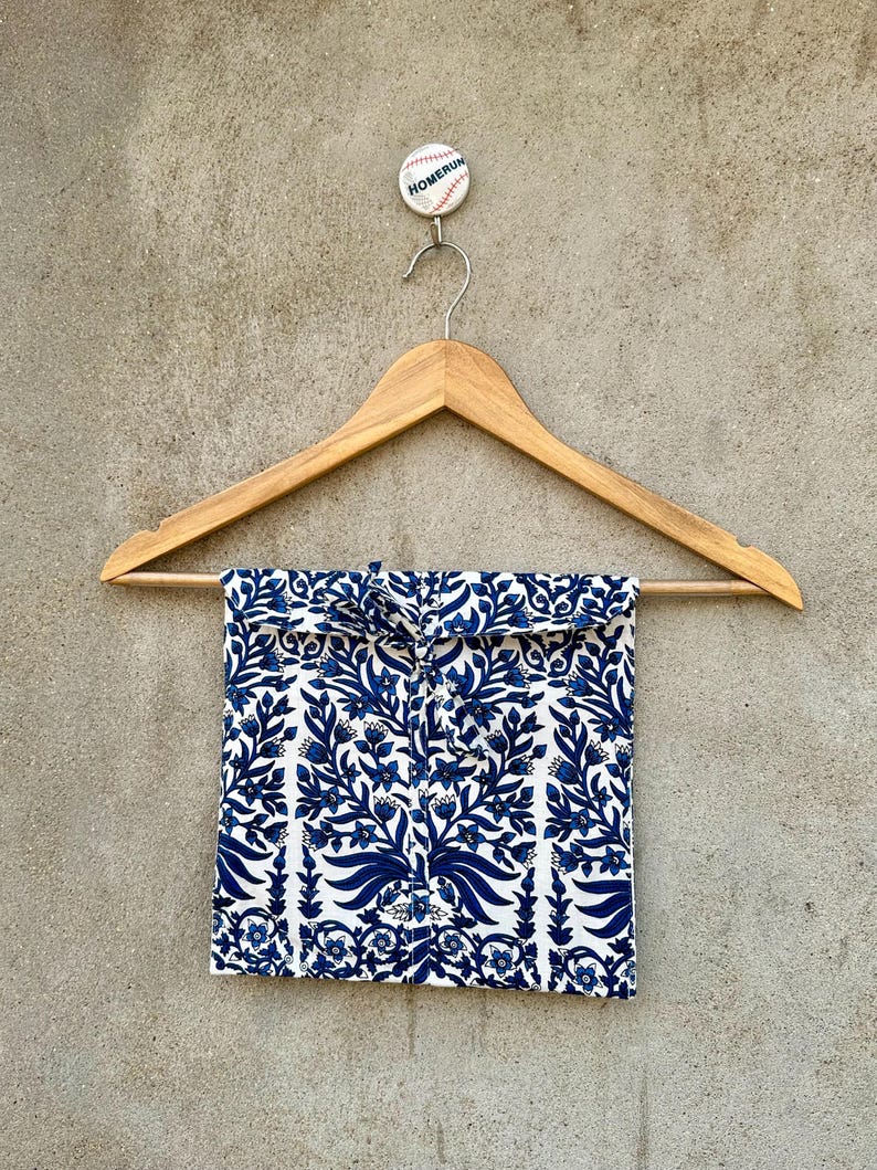 May include: A square fabric pouch featuring a blue and white floral pattern, suspended from a wooden hanger. The pouch has a tie closure at the top center. Above the hanger, a small circular pin displays the word HOMERUN in black text.