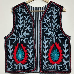 Könnte beinhalten: Eine schwarze ärmellose Weste mit hellblauen gestickten Blumenrankenmustern und roten Zierborten. Die Vorderteile zeigen ein zentrales rotes und türkisfarbenes Blumenmotiv. Das Innenfutter ist sichtbar und weist ein schwarz-weiß gestreiftes Muster auf.