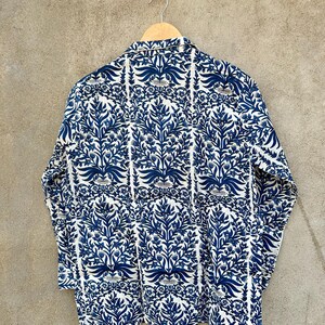May include: A long-sleeved button-down shirt featuring a repeating dark blue floral and botanical pattern on a white background. The shirt has a classic collar and is displayed on a wooden hanger, with a small baseball-themed pin attached above.