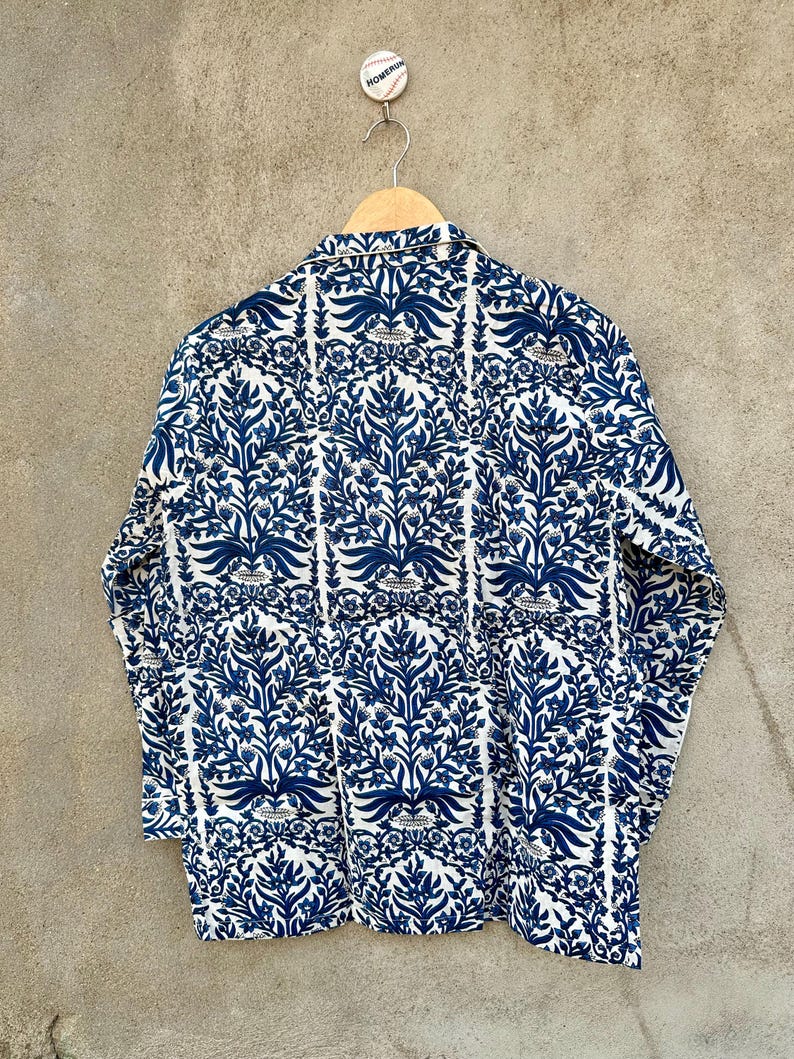 May include: A long-sleeved button-down shirt featuring a repeating dark blue floral and botanical pattern on a white background. The shirt has a classic collar and is displayed on a wooden hanger, with a small baseball-themed pin attached above.