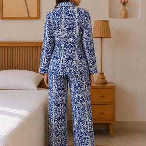 May include: A two-piece pajama set featuring a long-sleeved button-down shirt and matching wide-leg trousers. The fabric is white with a dense, intricate blue floral and botanical pattern, finished with white piping along the collar and cuffs.