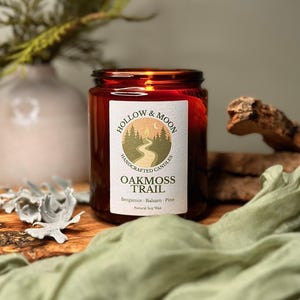 May include: A brown glass jar candle with a white label that reads "OAKMOSS TRAIL" and "HOLLOW & MOON HANDCRAFTED CANDLES". The candle is surrounded by greenery and a light green cloth. The candle's scent is bergamot, balsam, and pine.