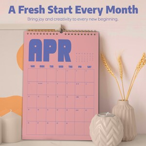 May include: A pink wall calendar for April with blue lettering and a gold spiral binding. The calendar has the text "A Fresh Start Every Month" and "Bring joy and creativity to every new beginning."